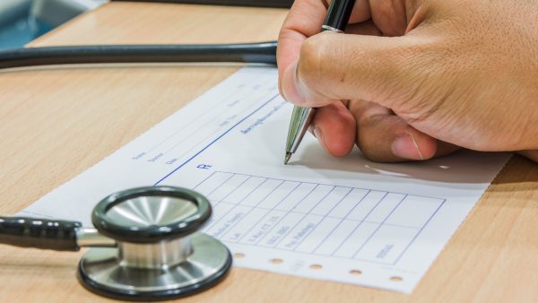 Mano escribiendo en un formulario médico junto a un estetoscopio.